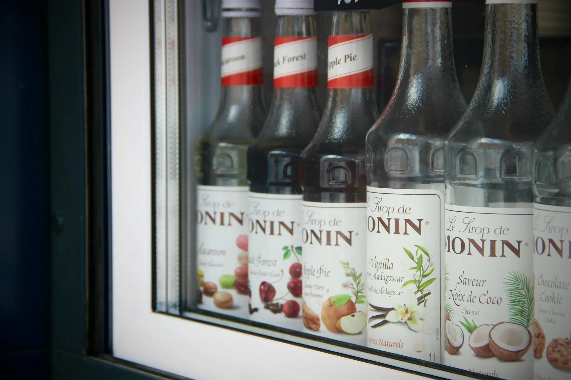 Close-up of Monin syrup bottles displayed in a bar window showcasing flavors like apple pie and coconut.