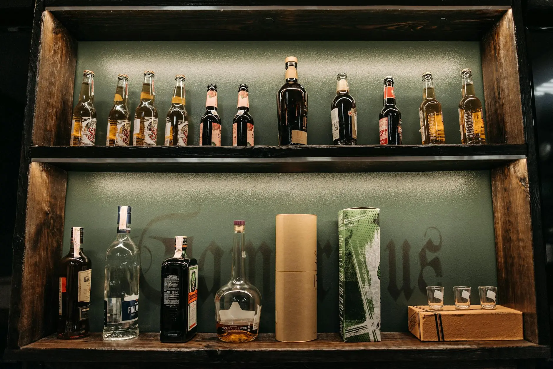 Rustic bar shelves displaying a variety of alcohol bottles, including beer and spirits, with shot glasses.