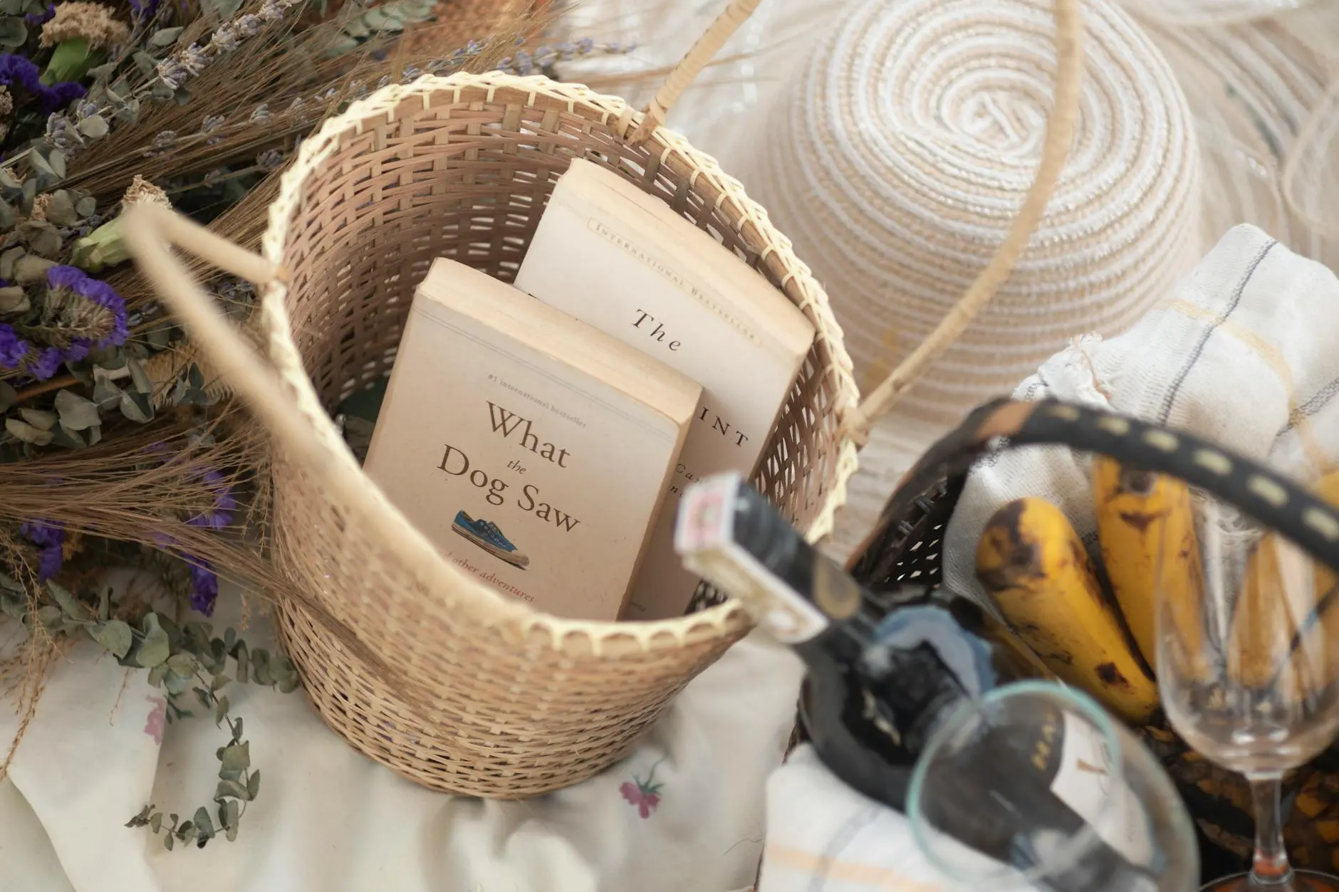 Top view of a wicker basket with books, bananas, wine, and glasses, perfect for a relaxed picnic.